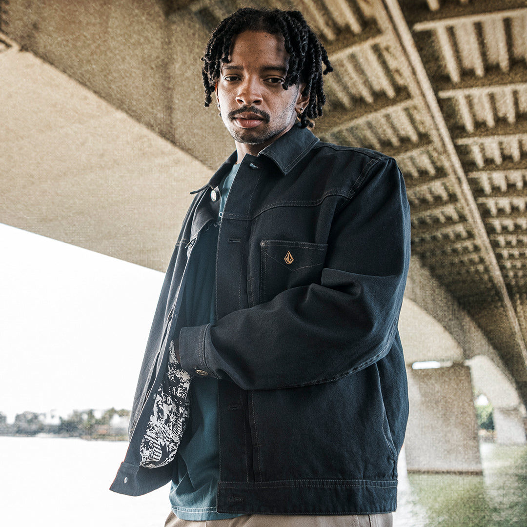 A man wearing a dark washed denim jacket over a teal shirt, standing under a concrete bridge. The jacket features a small gold diamond logo on the chest pocket.