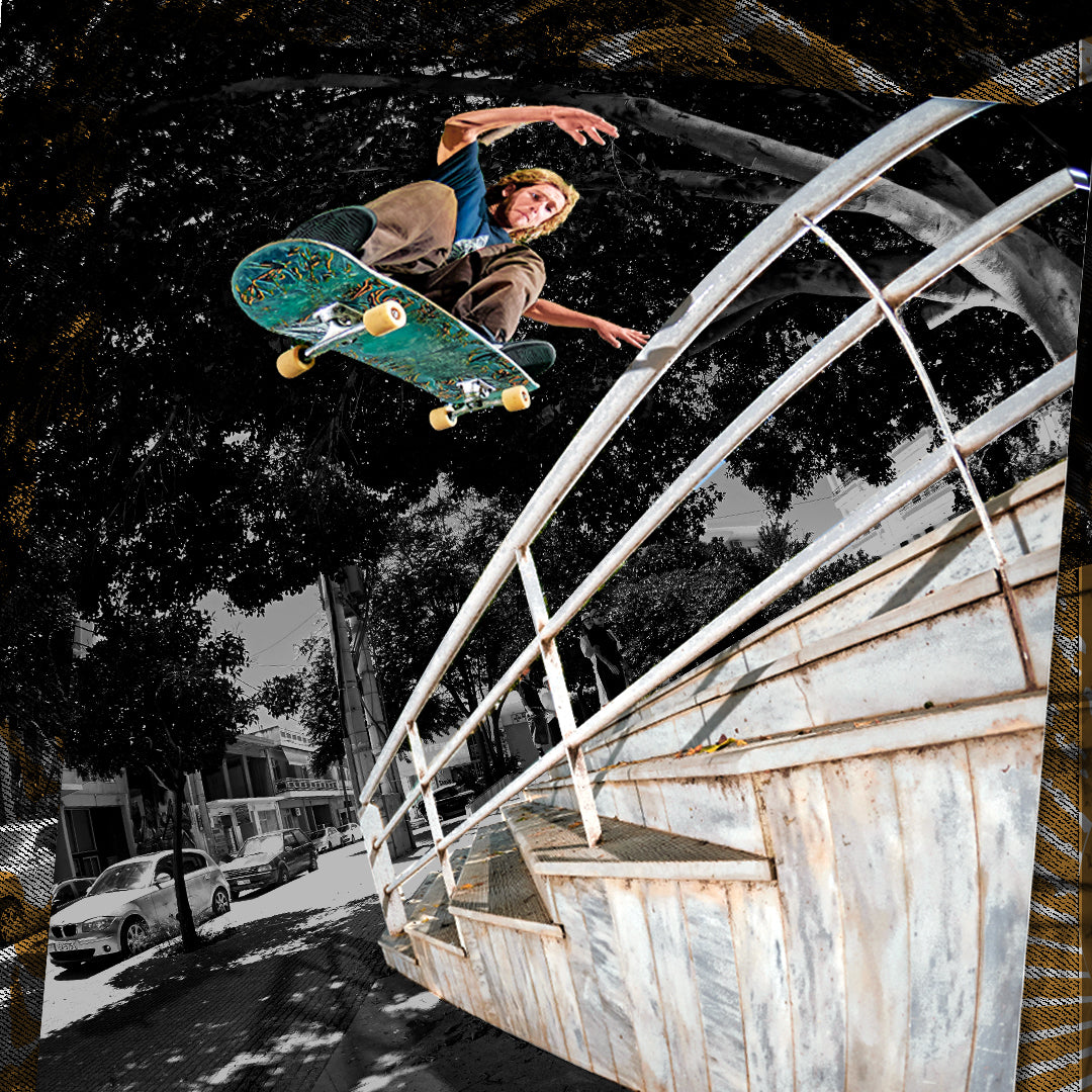 A skateboarder mid-air performing a trick above a metal stair rail in an outdoor urban setting, with trees and parked cars visible in the background. The skateboarder and skateboard are highlighted against a desaturated black-and-white scene.