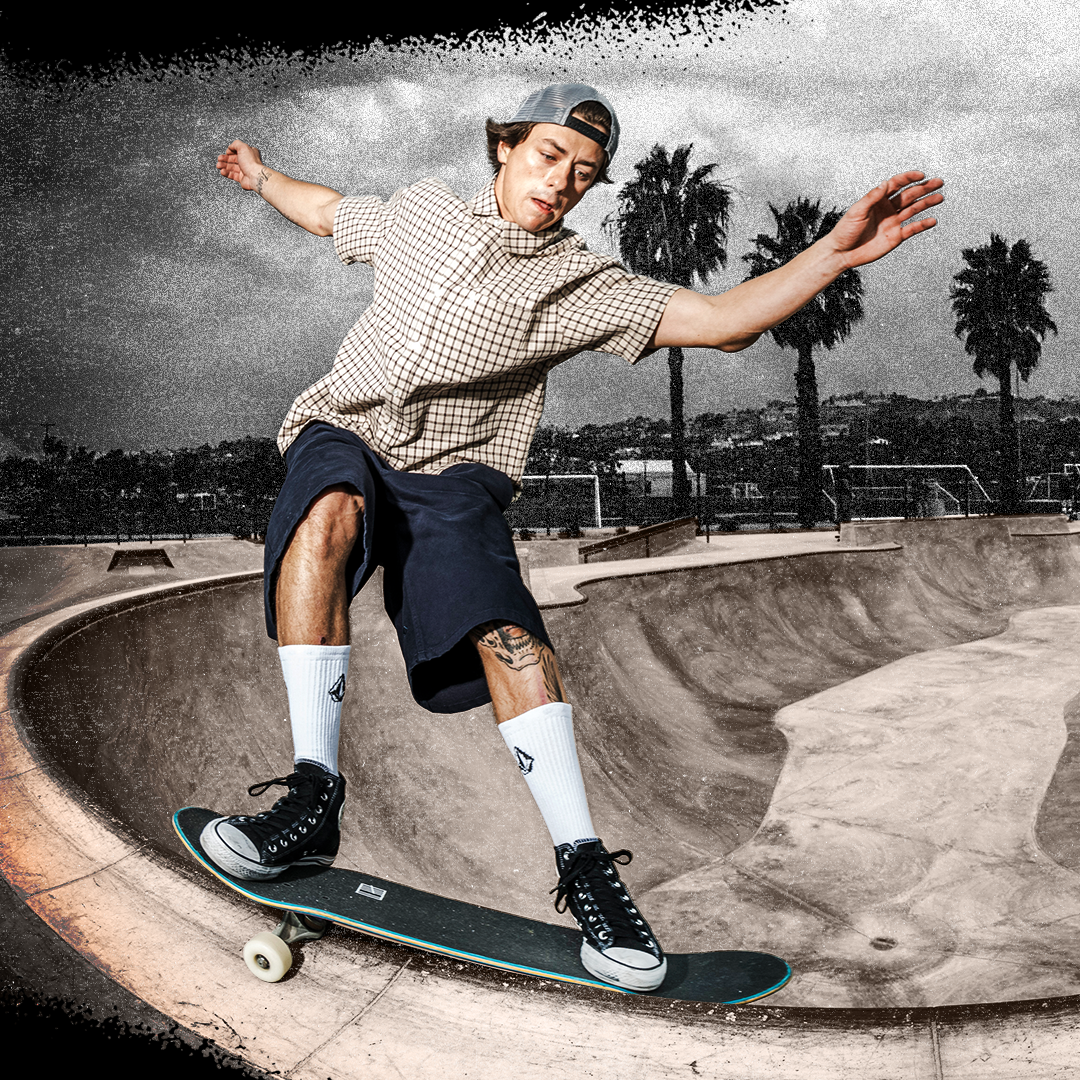 A skateboarder in a checkered shirt, dark shorts, white socks, and black high-top sneakers performs a trick on a skateboard inside a concrete skatepark bowl, with palm trees and a cloudy sky in the background.