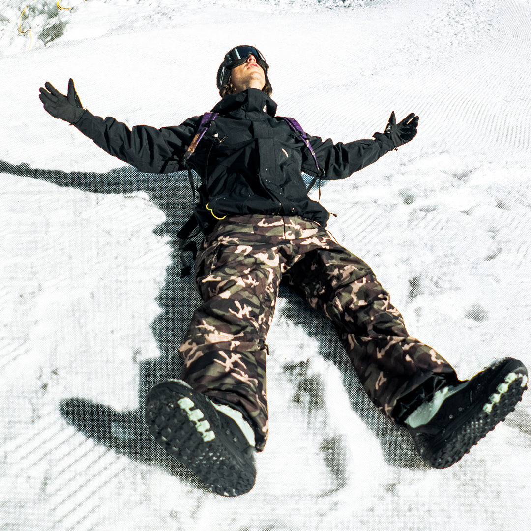 A person lying in the snow with arms outstretched, wearing a black ski jacket, camouflage snow pants, black gloves, and black ski boots.