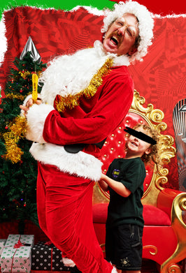 A man dressed as Santa Claus in a red suit with white fur trim and gold tinsel, holding a toy axe, posing next to a young child wearing a black blindfold and black shirt. They are set against a festive red background with a golden throne and a Christmas tree.