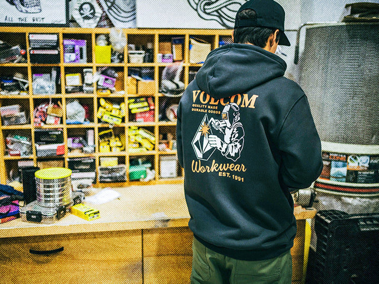 A person in a dark Volcom Workwear hoodie stands in a cluttered workshop with cubby shelves filled with tools and supplies. The hoodie features a graphic and text on the back.