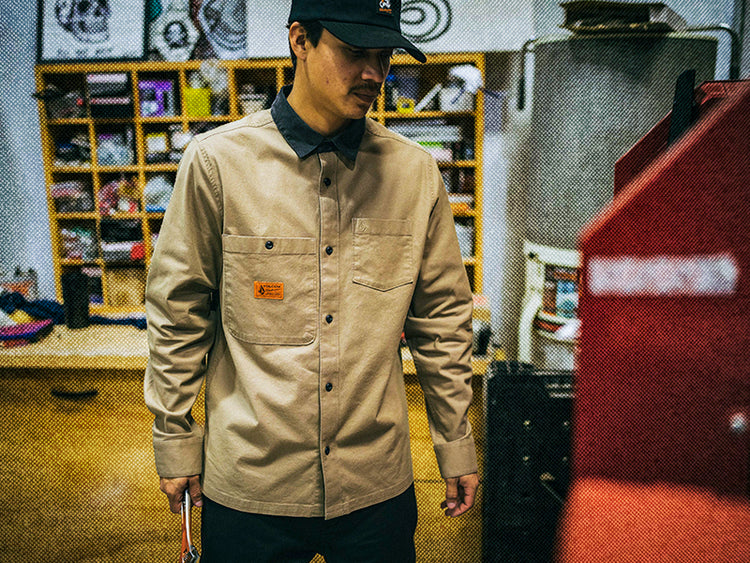 A person wearing a tan button-up work shirt and black cap stands indoors in a workshop, surrounded by shelves, tools, and equipment.