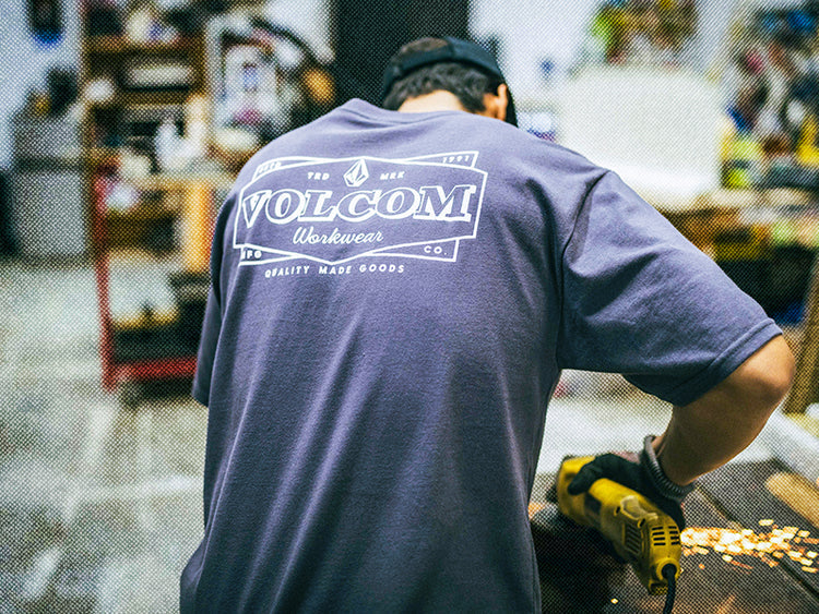 A person wearing a gray VOLCOM Workwear t-shirt is using a power tool, creating sparks, in a workshop filled with various tools and supplies. The image is taken from behind the person.