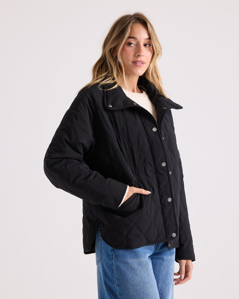 A woman wearing the Volcom Women’s Quinn Jacket—a black quilted shacket with snap buttons—paired with blue jeans stands against a plain light background, one hand in her pocket as she looks at the camera.