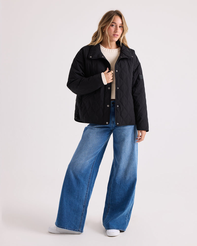 A person stands against a plain background, wearing the Volcom Women’s Quinn Jacket in black—a water resistant quilted style—over a cream sweater, wide-leg blue jeans, and white sneakers.
