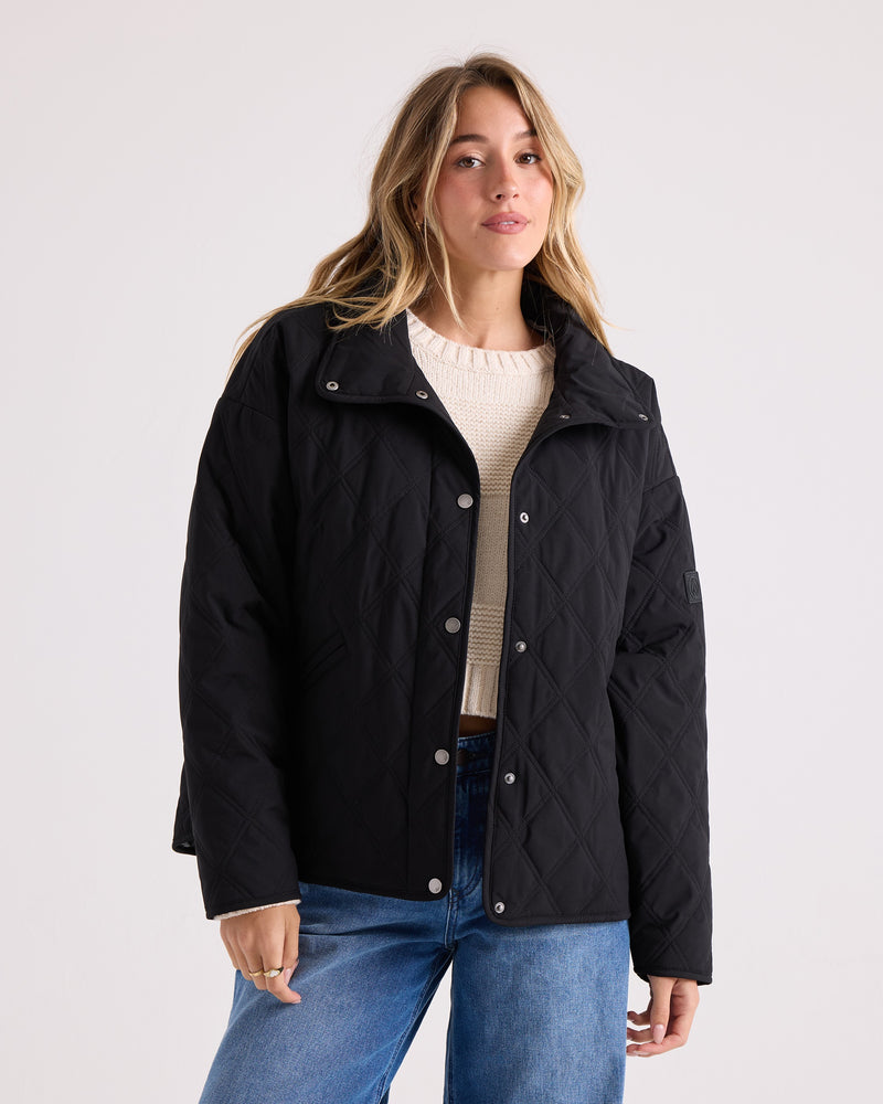 A woman wears the Volcom Women’s Quinn Jacket—a black quilted jacket with a quilted hood—layered over a cream sweater and blue jeans, standing against a plain light background and looking at the camera with a neutral expression.