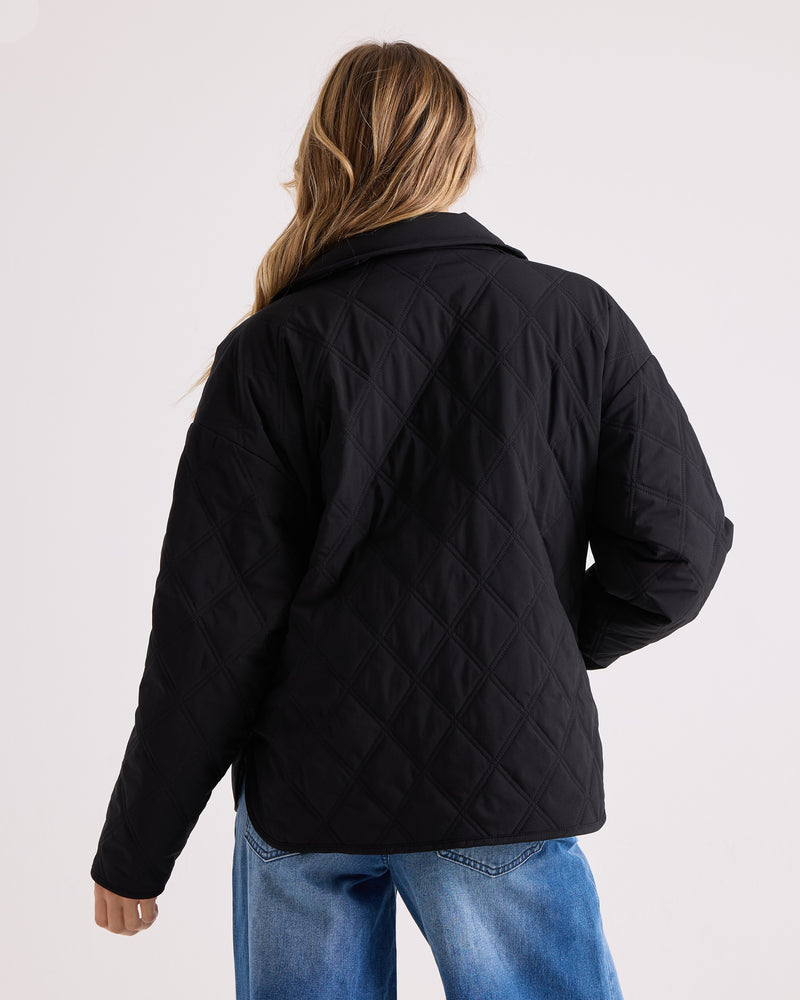 A person with long, wavy hair, seen from behind, wears the Volcom Women’s Quinn Jacket—a black quilted shacket with a water resistant finish—paired with blue jeans. The background is plain and light-colored.