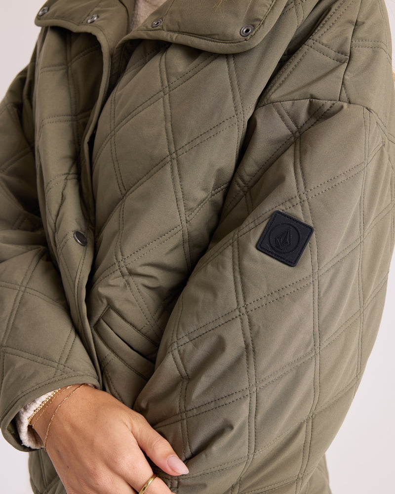Close-up of a person wearing the Volcom Women’s Quinn Jacket—an olive green quilted, hooded shacket featuring a black patch on the sleeve. With arms crossed, hands partially visible adorned with gold rings and a delicate bracelet, this water-resistant and wind-resistant jacket perfectly blends style and function for modern layering.