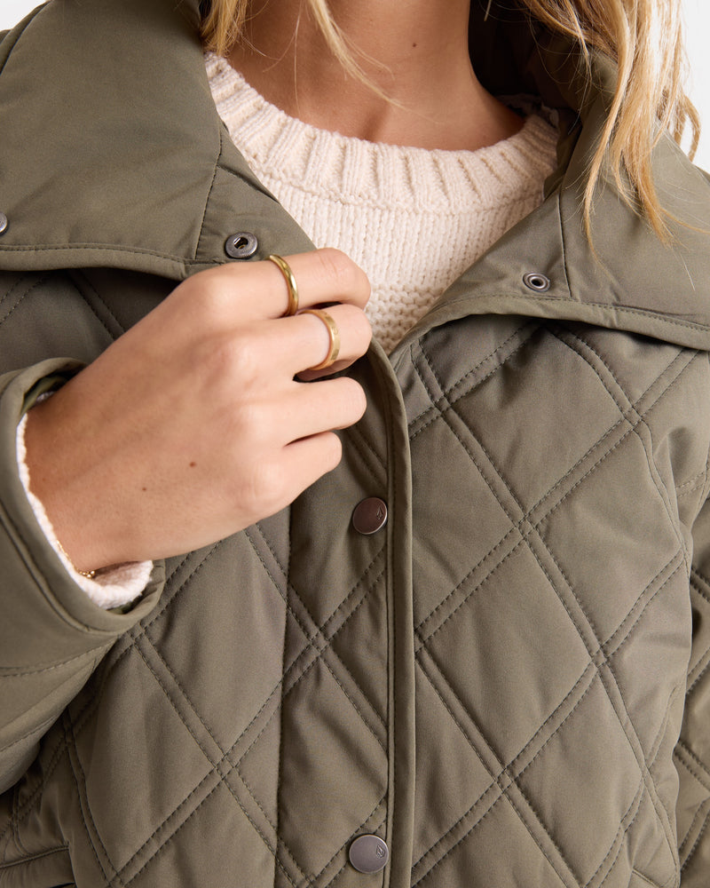 A person wearing the Volcom Women’s Quinn Jacket—an olive green, quilted, hooded shacket—over a cream knit sweater, has their hand near the collar displaying two gold rings. This water-resistant and wind-resistant layer blends warmth with effortless style.
