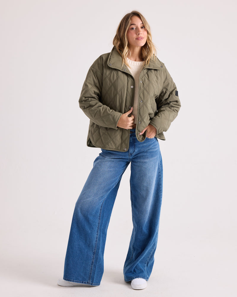 A woman with blonde hair stands against a plain background wearing the Volcom Women’s Quinn Jacket, styled in green with a quilted, hooded design. She pairs it with wide-leg blue jeans and white sneakers while holding the jacket’s collar. The wind-resistant material of the jacket makes this look both stylish and practical for cooler days.