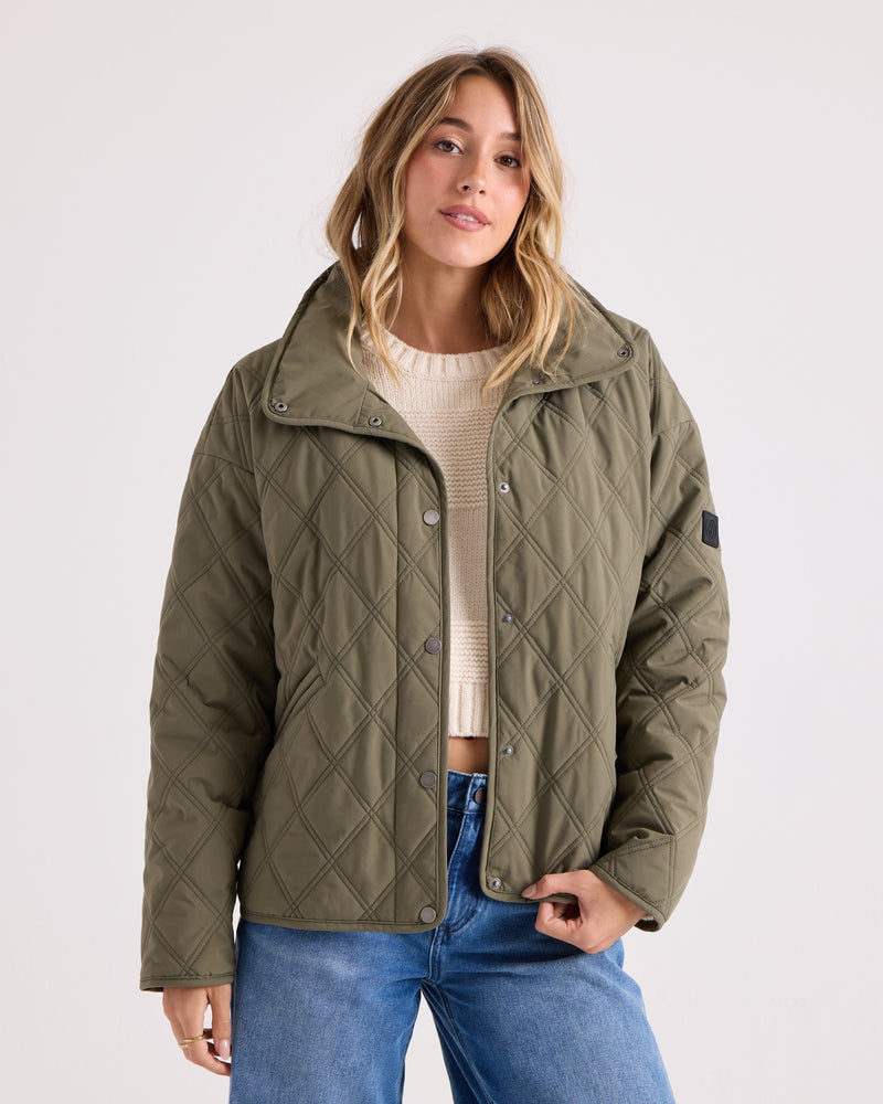 A woman stands against a plain light background, looking at the camera with a relaxed expression. She’s wearing the Volcom Women’s Quinn Jacket in olive green over a cream sweater and blue jeans, seamlessly blending laid-back style with practical comfort thanks to its wind resistant material.
