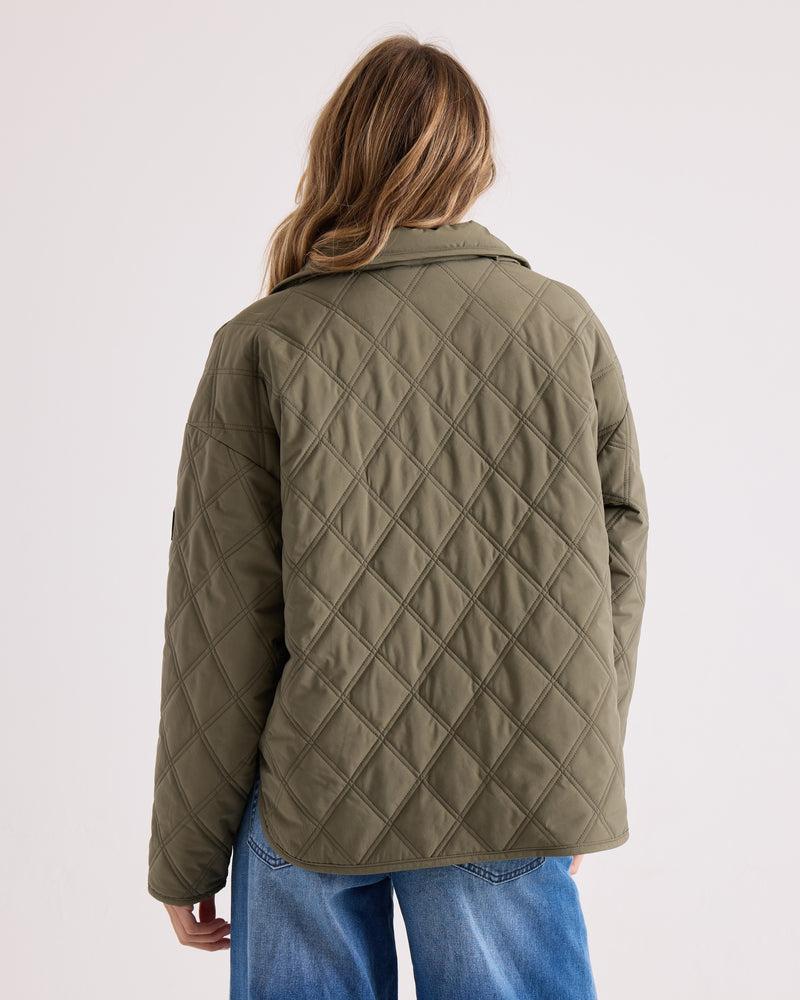 A person is seen from behind against a plain light background, wearing the Volcom Women’s Quinn Jacket in olive green paired with blue jeans.