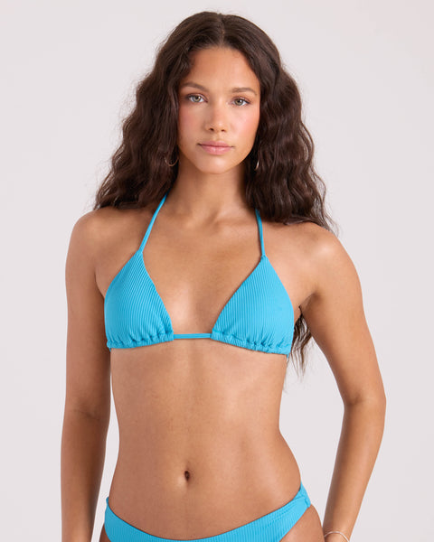 A woman with long, wavy brown hair wears the Volcom Women's Simply Rib Triangle Top in light blue, made from recycled nylon, standing against a plain light background and looking directly at the camera with a neutral expression.