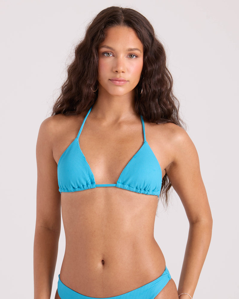 A woman with long, wavy brown hair wears the Volcom Women's Simply Rib Triangle Top in light blue, made from recycled nylon, standing against a plain light background and looking directly at the camera with a neutral expression.