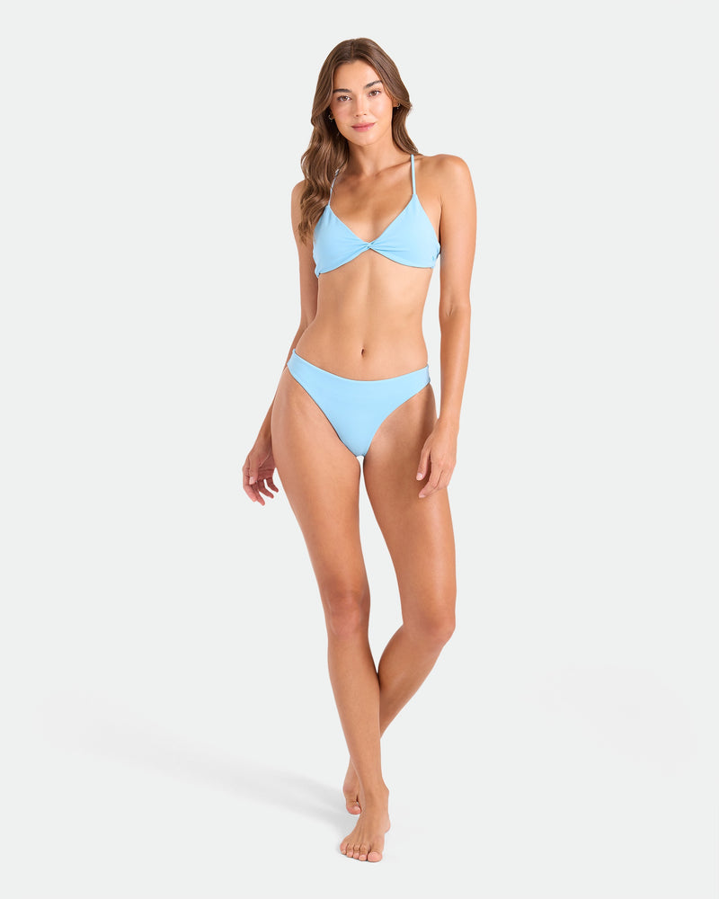 A woman with long brown hair and a neutral expression poses against a plain light background wearing the Volcom Women's Simply Seamless Twist Bralette in light blue, made from recycled nylon. One hand rests by her side, the other on her thigh.