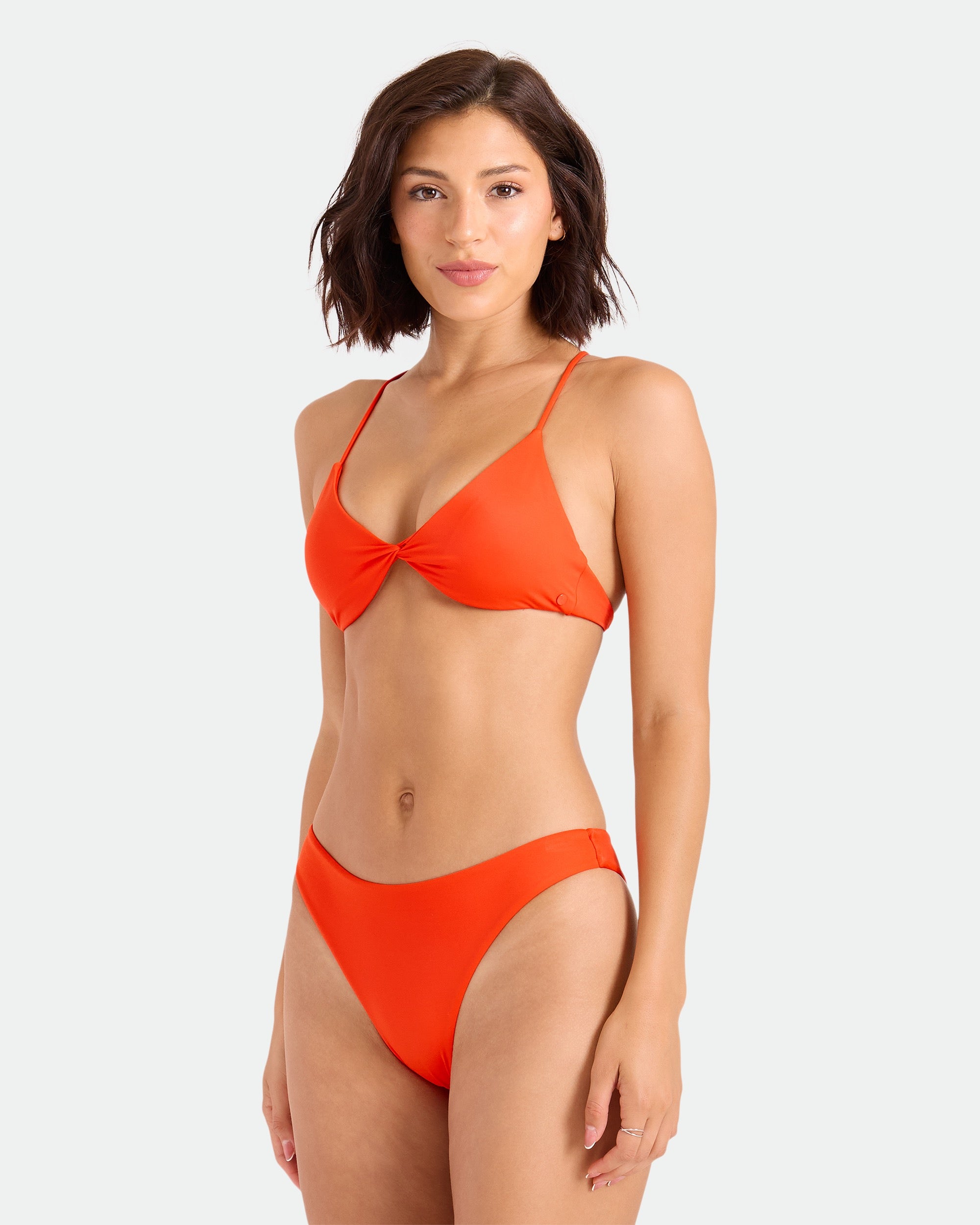 A woman with short brown hair wears the Volcom Women's Simply Seamless Twist Bralette in bright orange, made from recycled nylon, standing against a plain light background and looking at the camera with a slight smile.