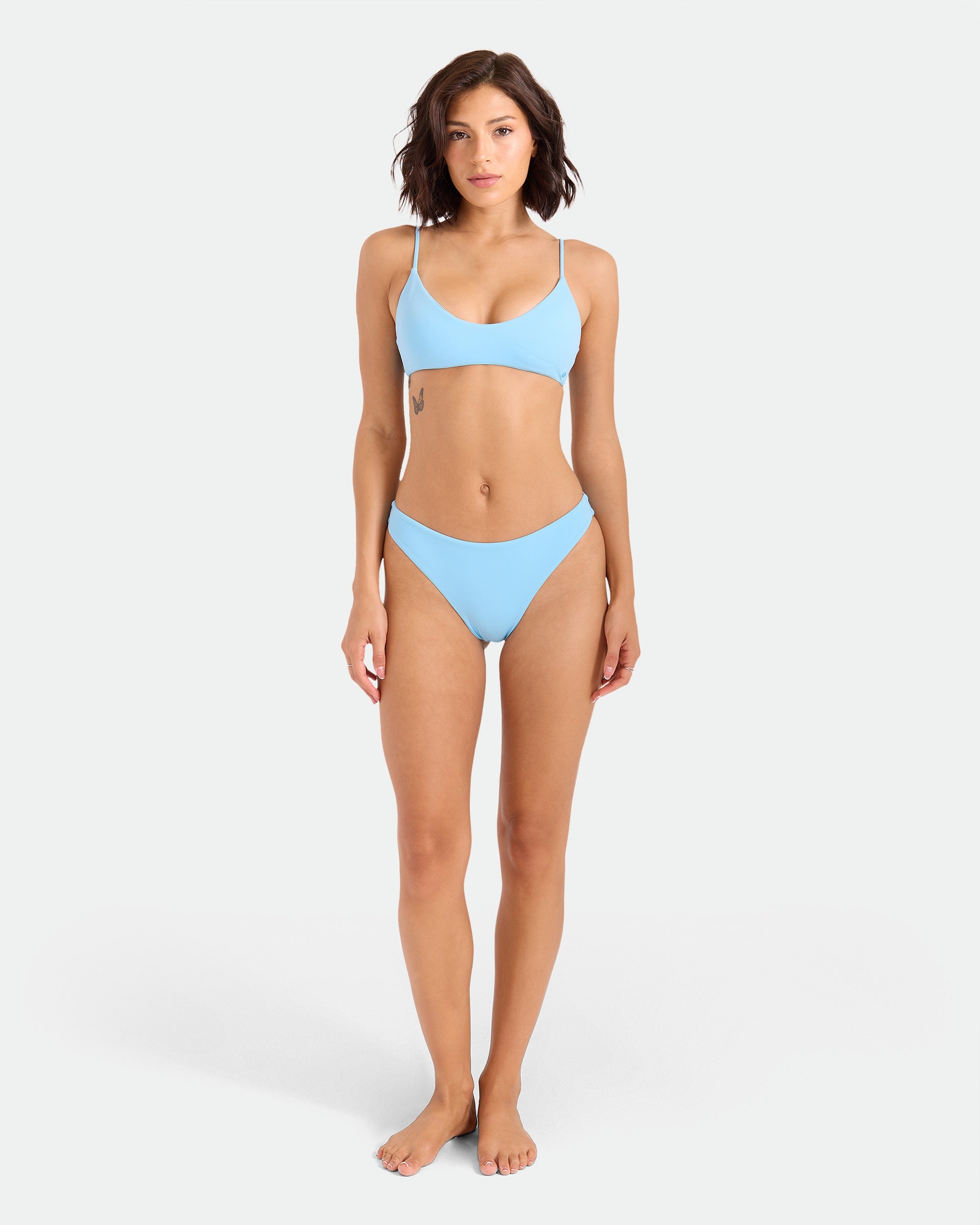 A woman with short brown hair stands barefoot against a white background, wearing the Volcom Women's Simply Seamless Midrise Skimpy bikini in light blue, featuring midrise fit and cheeky coverage made from recycled nylon.