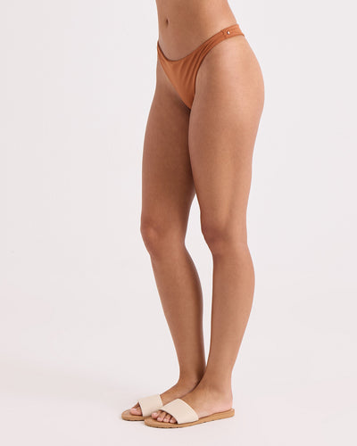 A person stands sideways against a plain light background, wearing Volcom's Women's Simply Seamless Scoop Tiny brown recycled nylon hipster swim bottom and beige sandals. Their head and upper body are not visible in the frame.