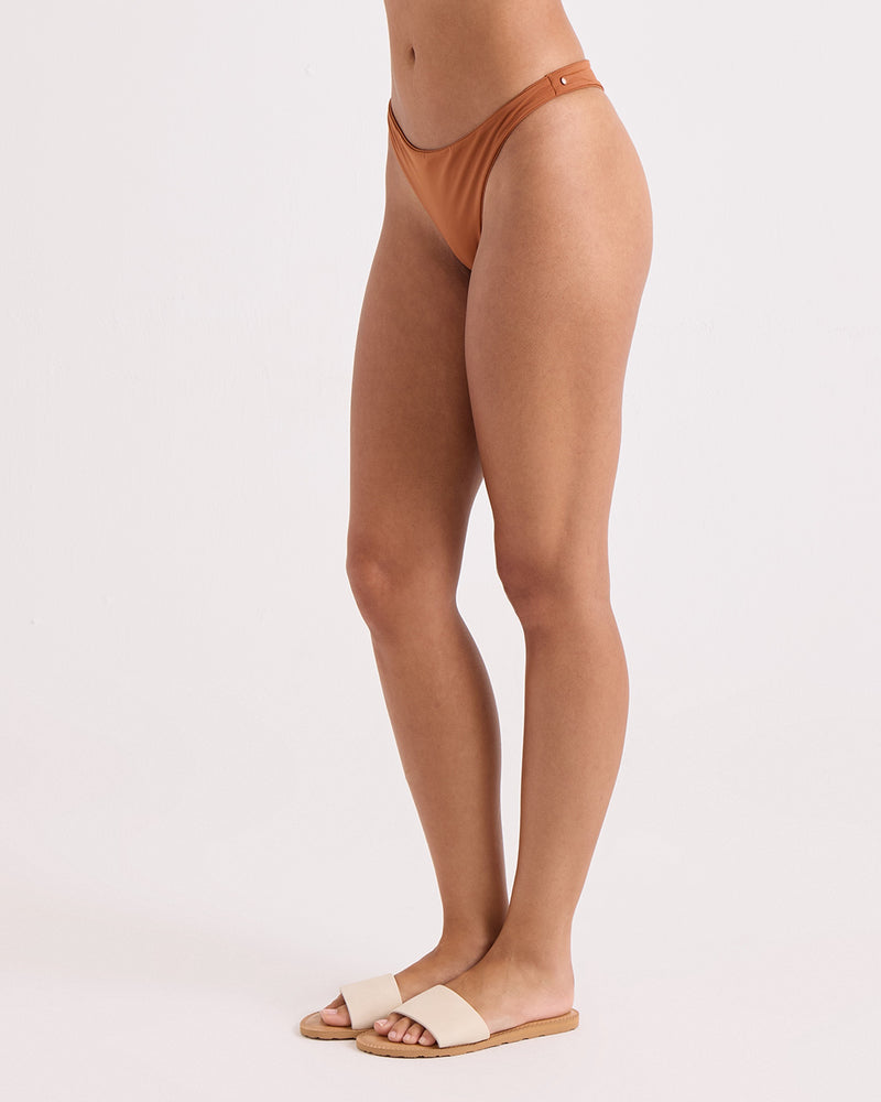 A person stands sideways against a plain light background, wearing Volcom's Women's Simply Seamless Scoop Tiny brown recycled nylon hipster swim bottom and beige sandals. Their head and upper body are not visible in the frame.