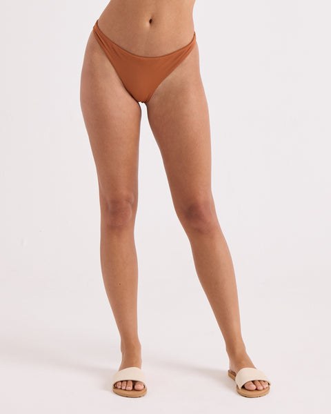A woman wears the Volcom Women's Simply Seamless Scoop Tiny hipster swim bottom, made from recycled nylon, and beige slide sandals. She stands against a plain white background with only her lower body visible.