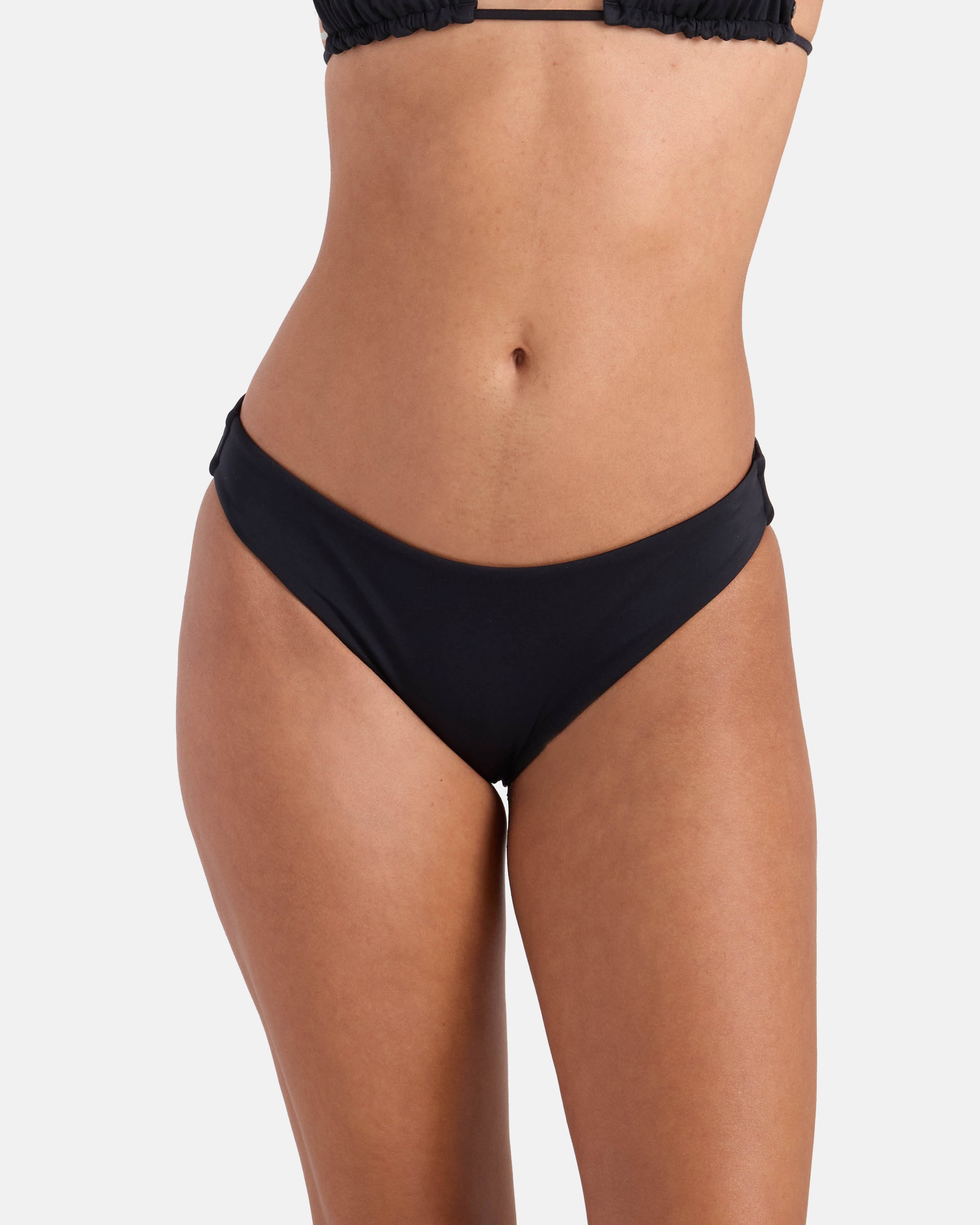 A woman models the Volcom Women's Simply Seamless Hipster Full black bikini bottom and matching top, made from recycled nylon, shown midriff to upper thighs against a plain white background.