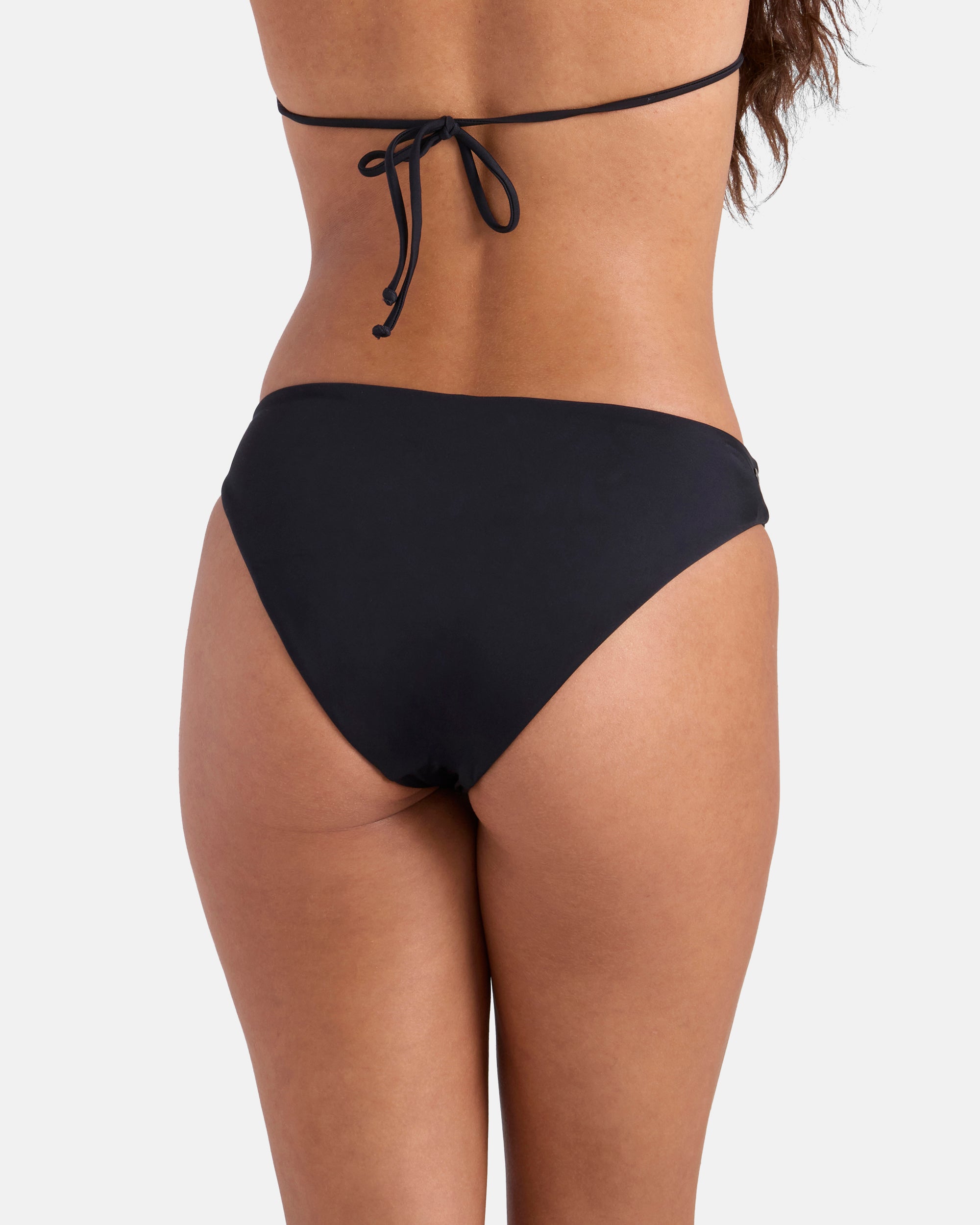 A woman is seen from the back wearing Volcom's Women's Simply Seamless Hipster Full black bikini bottom and a matching string-tied black bikini top, both made from recycled nylon, against a plain white background.