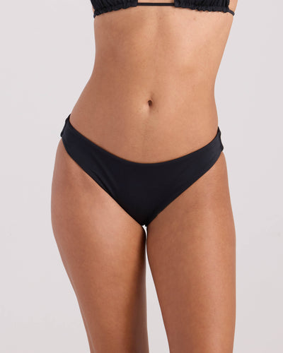 A woman models the Volcom Women's Simply Seamless Hipster Full, a black swimwear set with a hipster bikini bottom and matching top, shown from mid-torso to upper thighs against a plain light background.