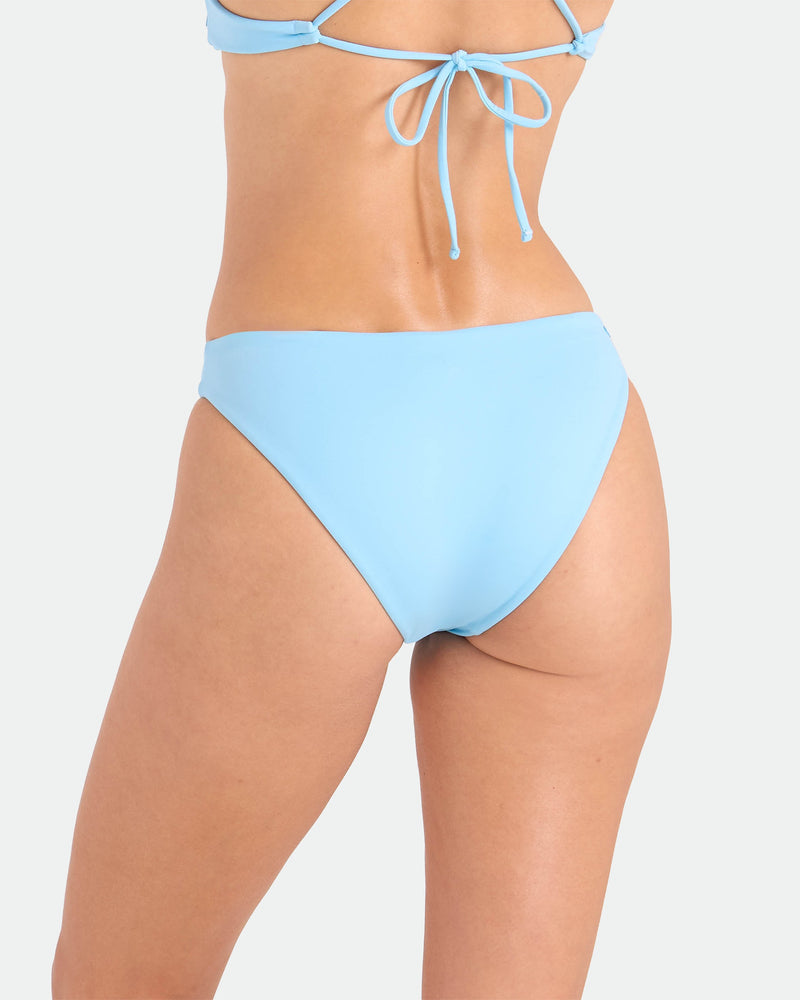 A person shown from the back wears Volcom's Women's Simply Seamless Hipster Full bikini in light blue, made from recycled nylon, with a back tie top and full coverage bottom, set against a plain white background.