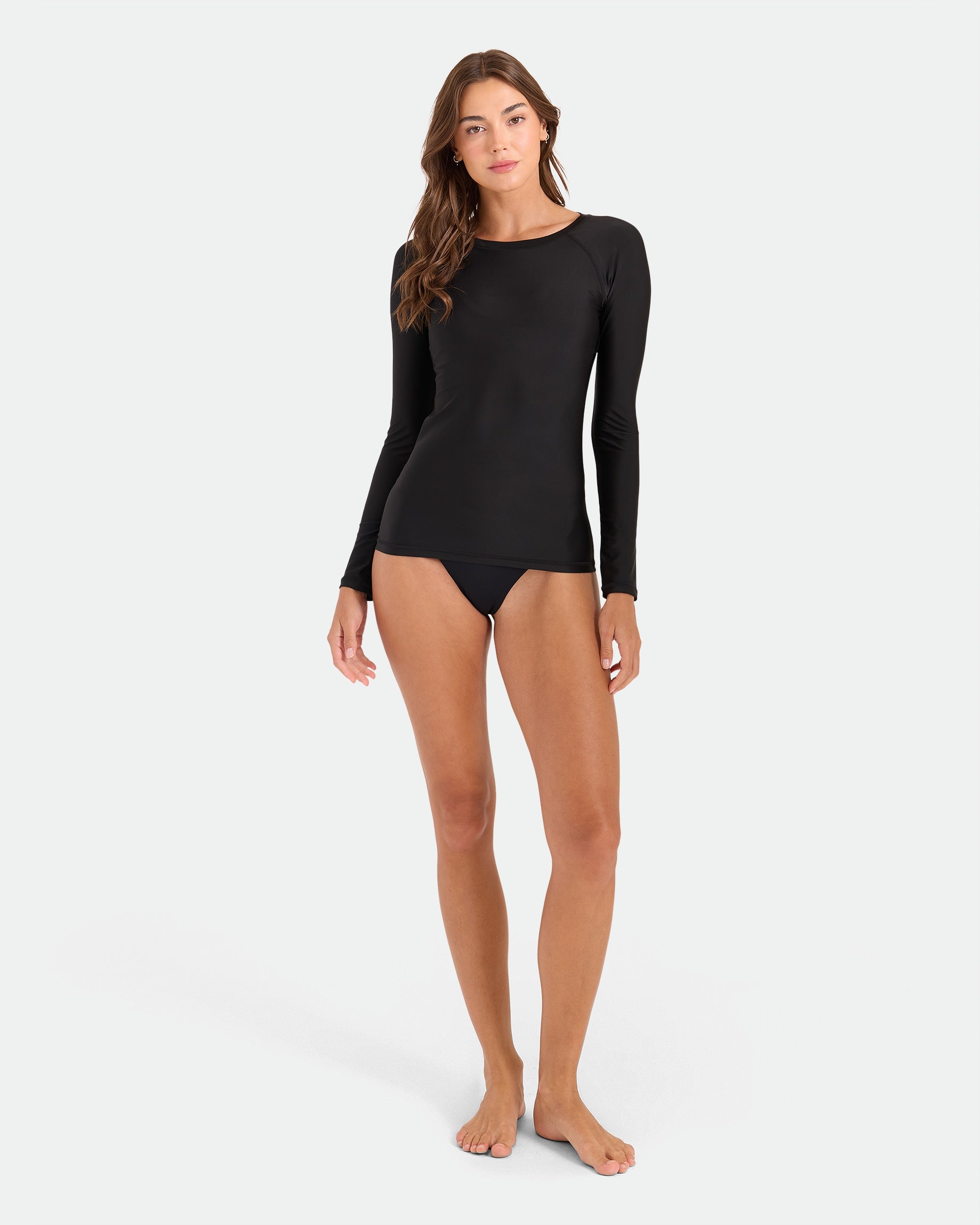 A woman stands barefoot against a plain background, wearing the Volcom Women's Simply Core Long Sleeve Rashguard and bikini bottoms. Her relaxed look highlights the easy pullover fit and all-day comfort of this fitted black swim top.