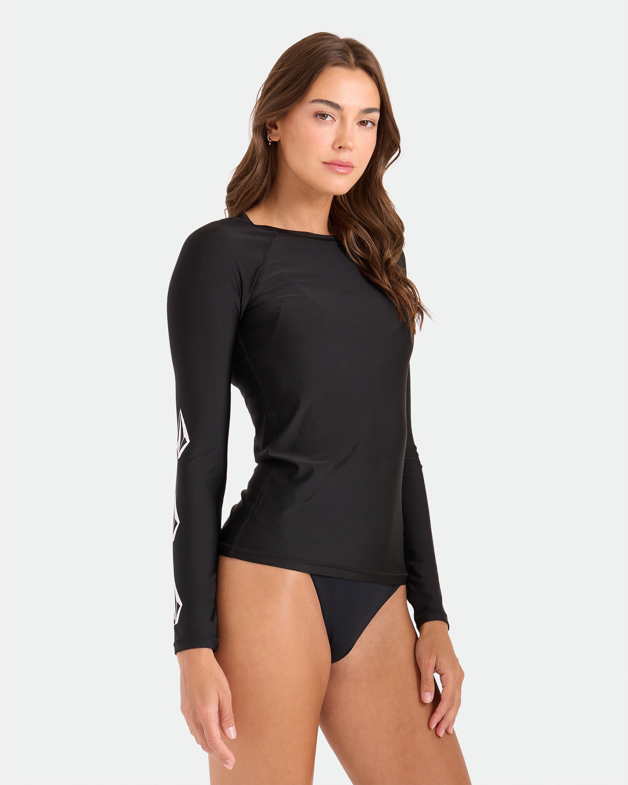 A woman stands against a plain light background wearing the Volcom Women's Simply Core Long Sleeve Rashguard with white graphic sleeve designs and matching black bikini bottoms, featuring a pullover fit for all-day comfort.