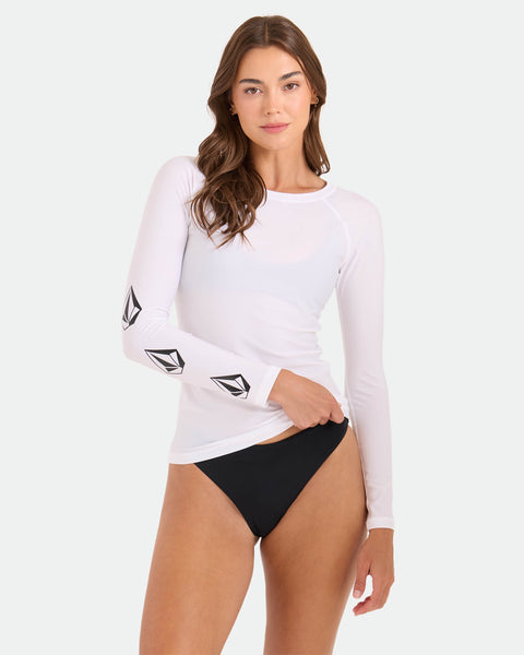 A woman models the Volcom Women's Simply Core Long Sleeve Rashguard, featuring black geometric sleeve logos, paired with black bikini bottoms. She stands against a plain light background, looking at the camera.