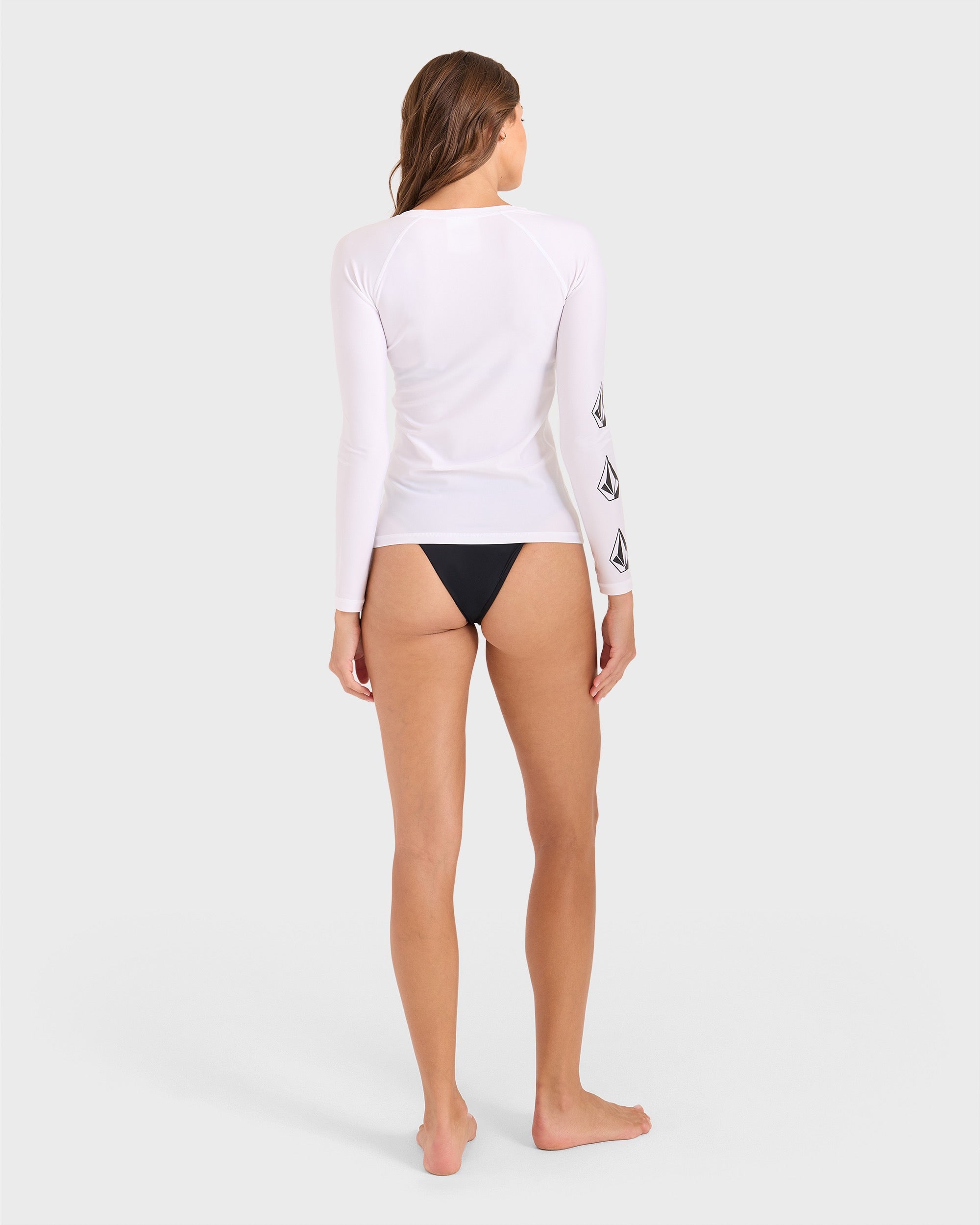 A woman stands barefoot facing away, wearing the Volcom Women's Simply Core Long Sleeve Rashguard with black logos on the left sleeve and black bikini bottoms. The background is plain light gray.