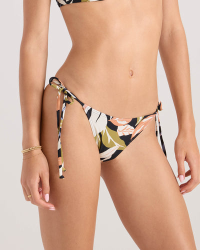 A woman wears the Volcom Women's Distant Tropic Tie Side Skimpy bikini bottom, featuring a floral print and crafted from recycled nylon. Shown mid-torso to upper thighs, she accessorizes with gold bracelets and a neutral manicure against a plain background.