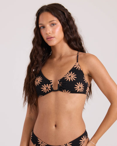 A woman with long wavy brown hair wears the Volcom Women's Bright & Sol Uwire Bralette, a black bikini top with tan floral patterns and thin straps, standing against a plain light background.