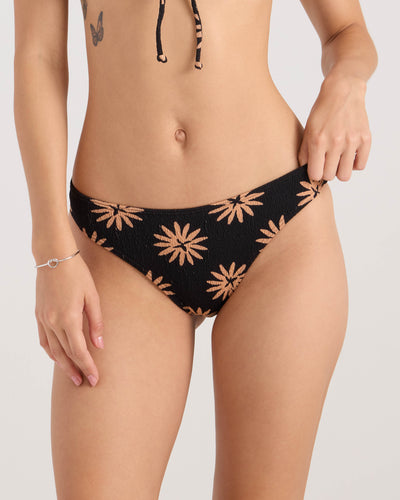 A woman, cropped from the shoulders down, wears the Volcom Women's Bright & Sol Midrise Skimpy bikini bottom with a tan floral pattern and high-cut silhouette. She has a tattoo and a thin bracelet, holding the side of the bikini.