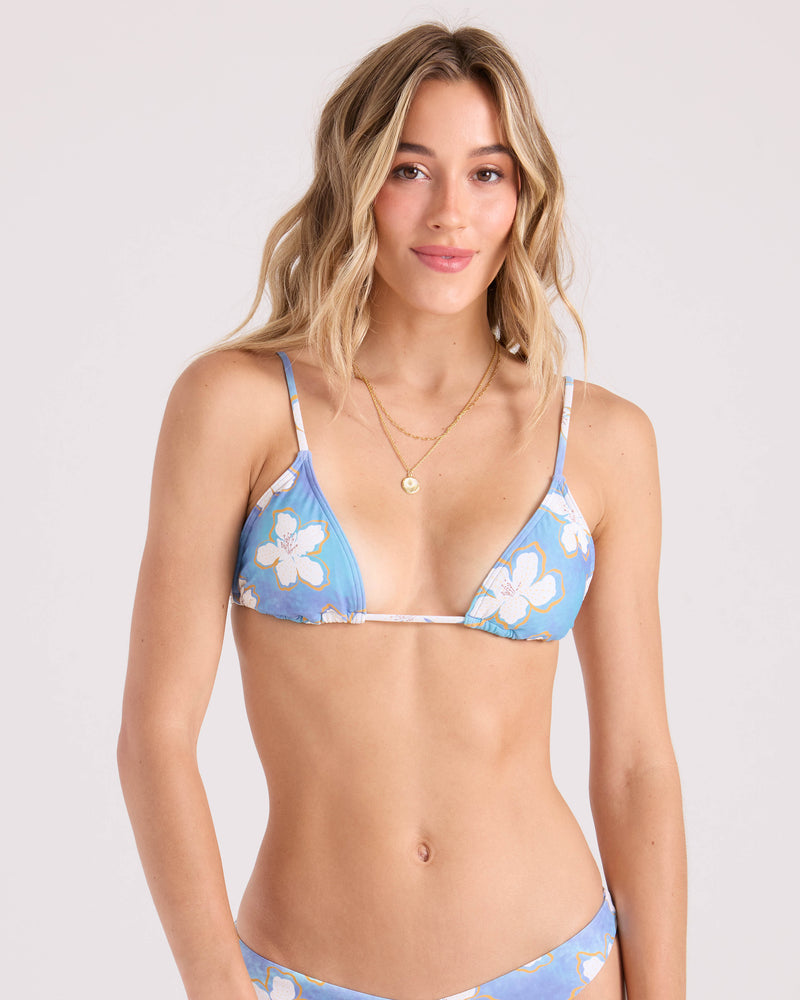 A woman with long, wavy blonde hair models the Volcom Women's Ocean Haze Triangle Top, a blue floral bikini with thin straps. She stands against a plain light background, smiling softly at the camera and wearing gold layered necklaces.