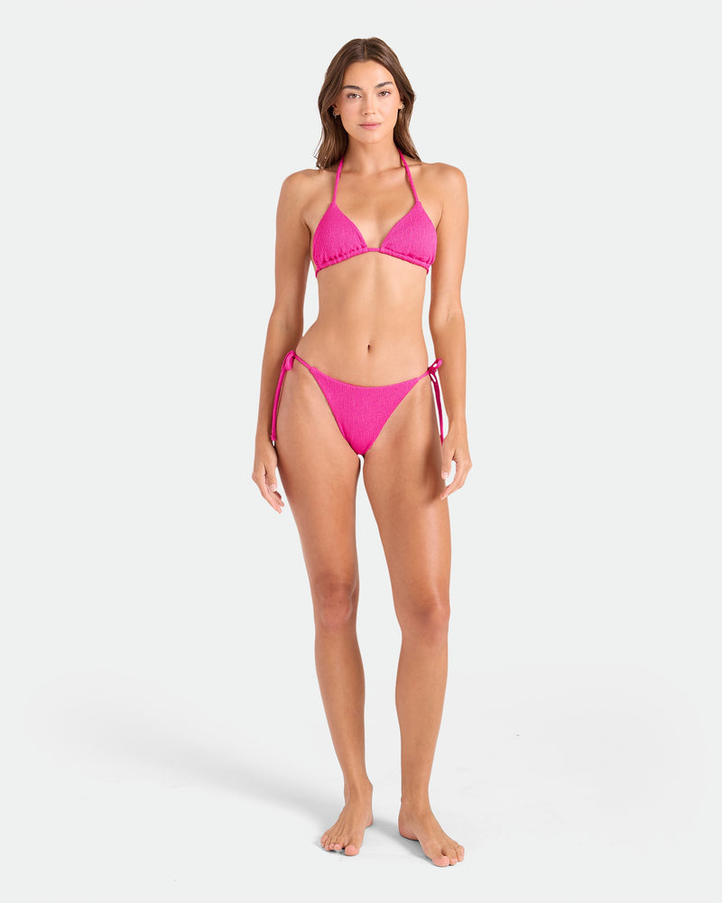 A woman with long brown hair and a neutral expression faces forward against a white background, wearing the Volcom Women's Pucker Up Tie Side Teeny Bikini Bottom in bright pink with adjustable ties and a hipster fit.