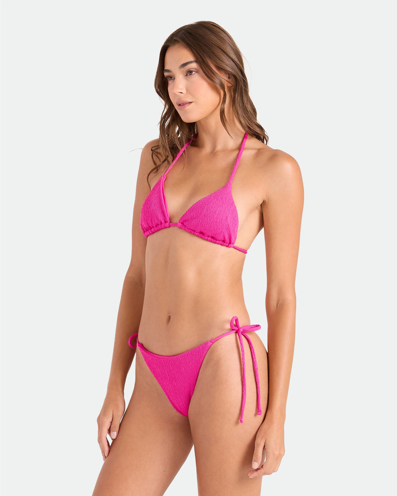 A woman with long wavy brown hair poses against a plain background wearing Volcom's Women's Pucker Up Tie Side Teeny Bikini Bottom in bright pink, featuring adjustable side ties. She looks slightly to the side.