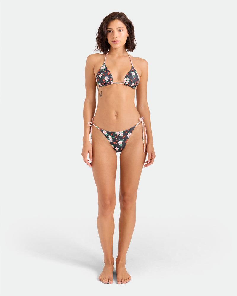 A woman faces forward against a plain light background, wearing the Volcom Women's Sweet & Spicy Reversible Triangle Top, a black bikini featuring a colorful floral pattern and adjustable tie straps.