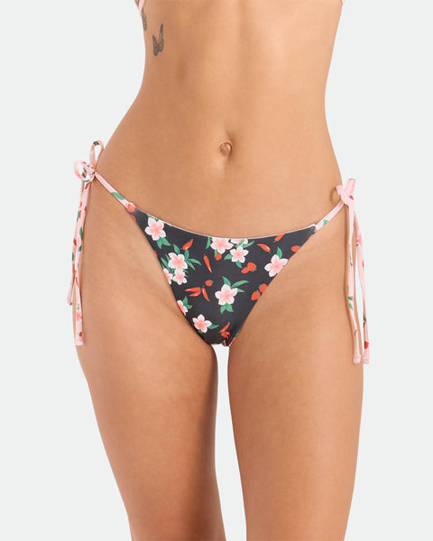 A model wears the Volcom Women's Sweet & Spicy Tie Side Teeny Reversible Bikini Bottom, featuring adjustable tie sides and a floral pattern. Only the torso and upper thighs are shown, with a small tattoo visible near the left hip.