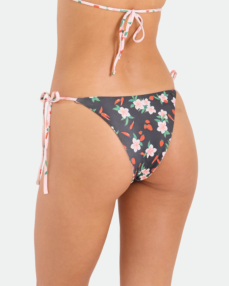 A woman wears the Volcom Women's Sweet & Spicy Tie Side Teeny Reversible Bikini Bottom, featuring cheeky coverage, adjustable tie sides, and a black base with pink and white florals. The photo focuses on her lower back and hip against a plain background.