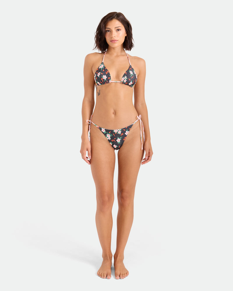 A woman faces forward, barefoot, in a Volcom Women’s Sweet & Spicy Tie Side Teeny Reversible Bikini Bottom with adjustable ties and cheeky coverage. She has short brown hair, a side tattoo, and stands against a plain white background.