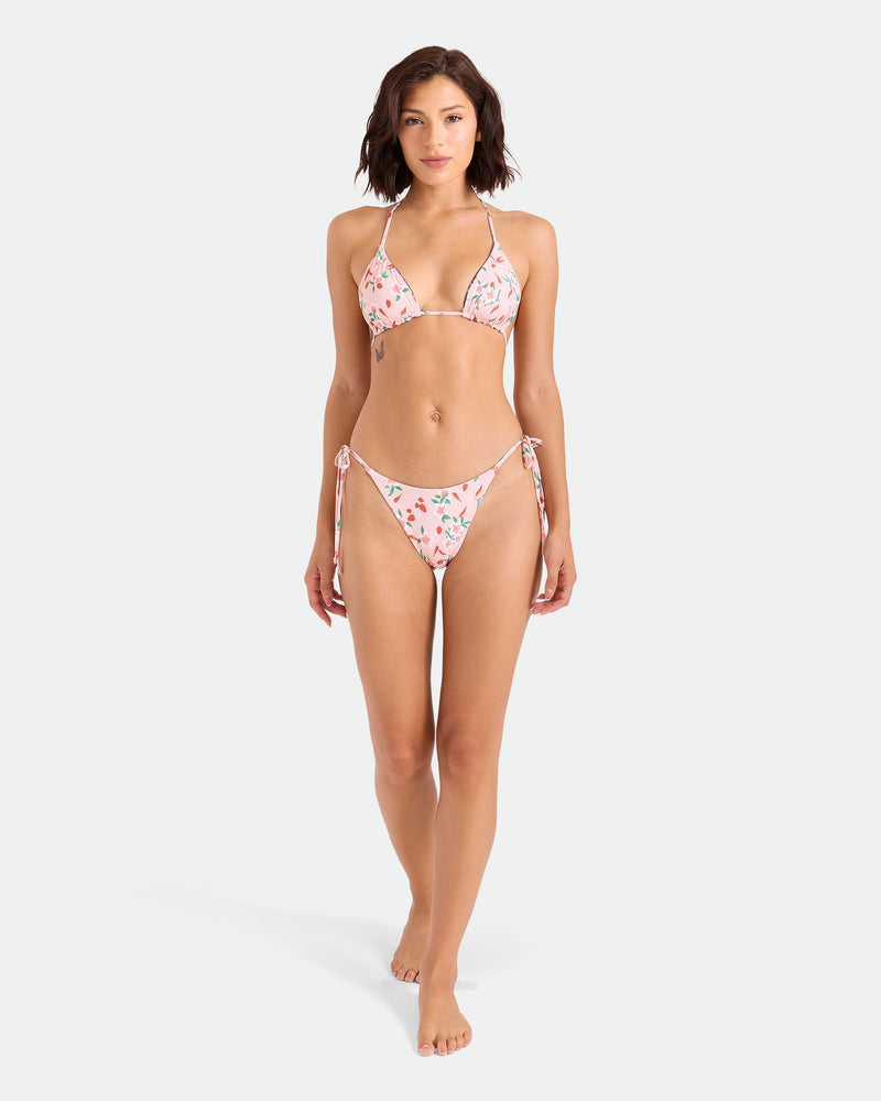 A woman with short brown hair stands barefoot against a white background, wearing the Volcom Women's Sweet & Spicy Tie Side Teeny Reversible Bikini Bottom featuring adjustable tie sides and cheeky coverage in a pink floral pattern.