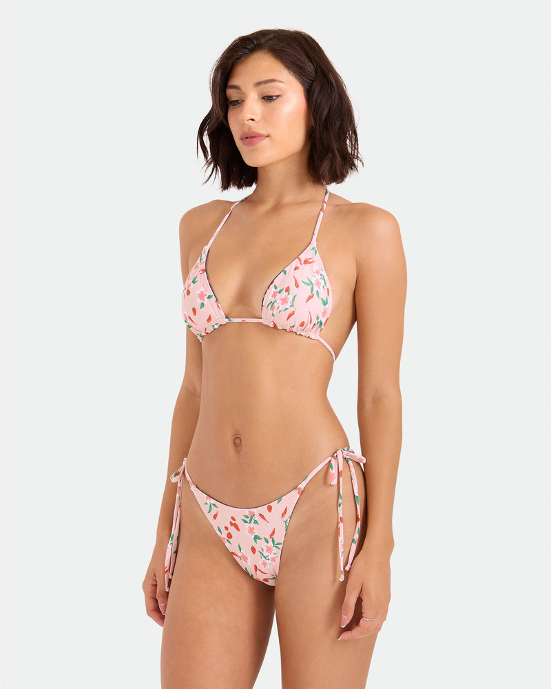 A woman with short brown hair models the Volcom Women's Sweet & Spicy Tie Side Teeny Reversible Bikini Bottom in a light pink cherry print. The cheeky bikini offers adjustable tie sides and triangle cups, set against a plain light gray background.