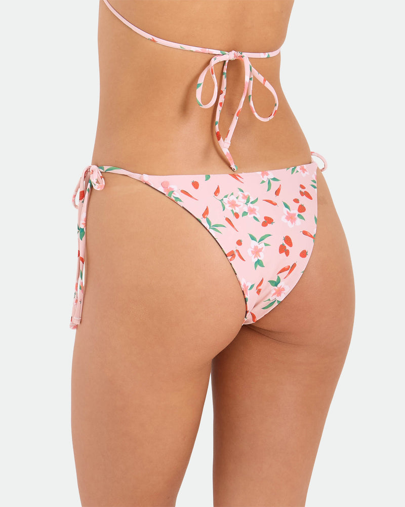 A person wears the Volcom Women's Sweet & Spicy Tie Side Teeny Reversible Bikini Bottom, featuring pink floral print, adjustable tie sides, and cheeky coverage, paired with a matching top tied at the back, highlighting the swimsuit's back design.