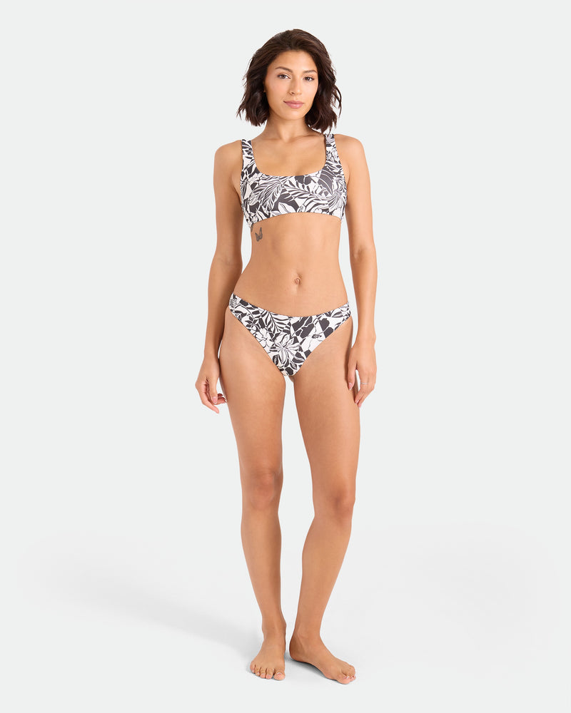 A woman stands barefoot against a plain light background, wearing the Volcom Women's Hibiscus Diamond Scoop Bralette—a black-and-white leaf-patterned bikini top made from recycled nylon. She faces forward with her arms relaxed by her sides.