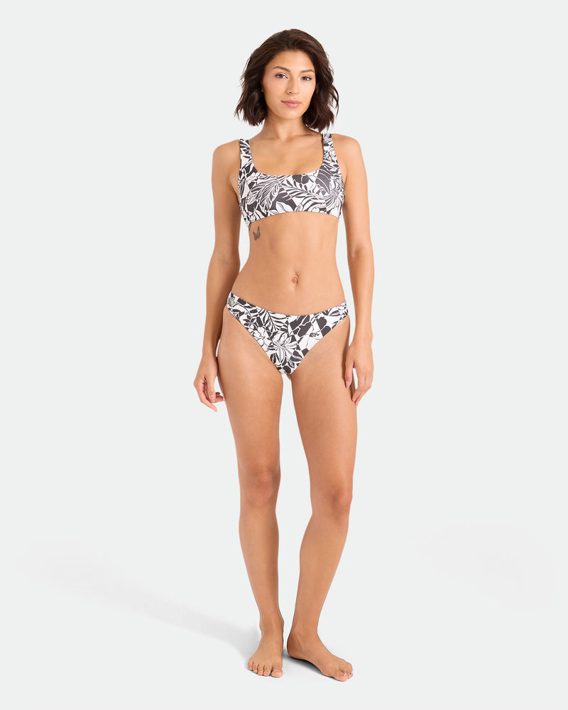A woman with shoulder-length dark hair stands barefoot against a white background, wearing Volcom's Women's Hibiscus Diamond Midrise Skimpy Reversible Bikini Bottom. She faces forward with a relaxed expression and neutral pose.