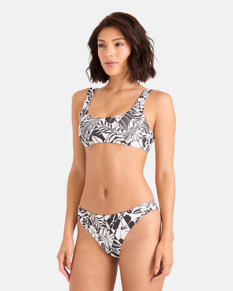 A woman with short brown hair wears the Volcom Women's Hibiscus Diamond Midrise Skimpy Reversible Bikini Bottom, featuring a black and white floral pattern, standing against a plain light background and looking slightly to the side.