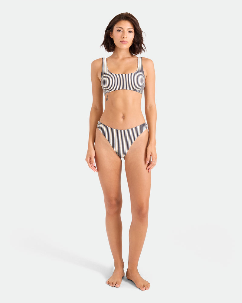 A woman stands barefoot against a white background, wearing the Volcom Women's Hibiscus Diamond Midrise Skimpy Reversible Bikini Bottom made from recycled nylon. She faces forward with a neutral expression and relaxed posture.